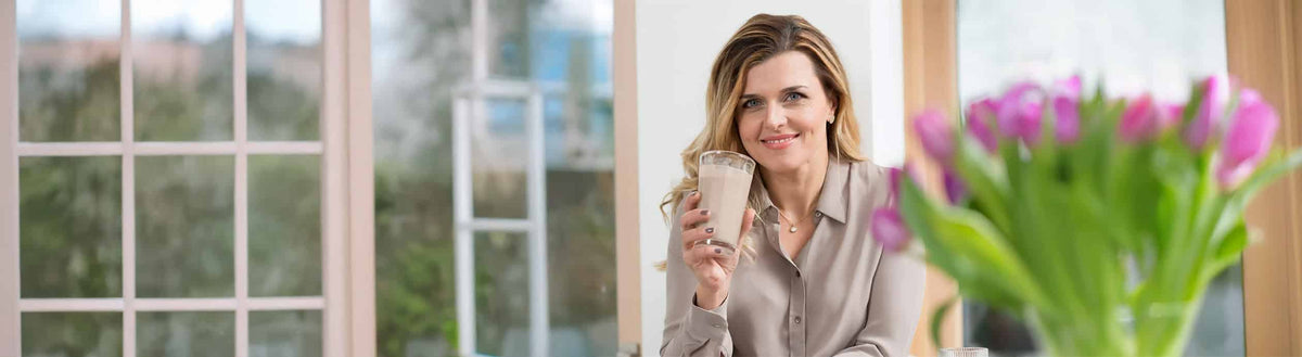 Lächelnde Frau hält einen Shake in der Hand, daneben ein Strauß pinker Tulpen