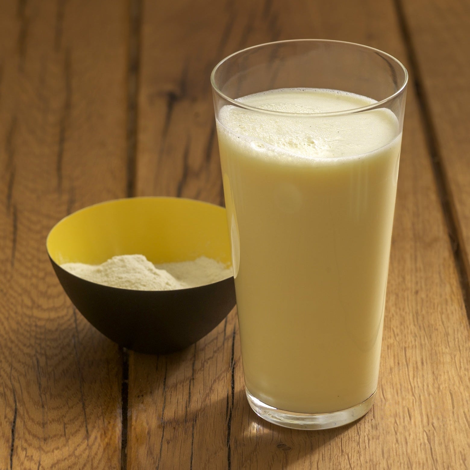 Ein Glas mit gelblicher Flüssigkeit steht neben einer schwarz-gelben Schale mit Milchpulver aus dem Precon 3-Monats-Mega-Vorrats-Paket CH, ideal für Precon Diät oder Gewichtsmanagement, auf einem Holztisch.