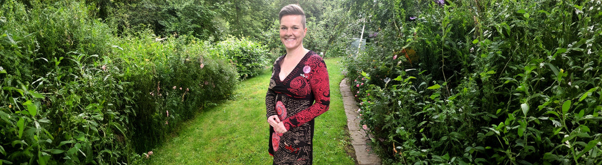 Julia (43) nach ihrer Gewichtsabnahme mit Precon im Kleid auf einem Gartenweg zwischen Grün