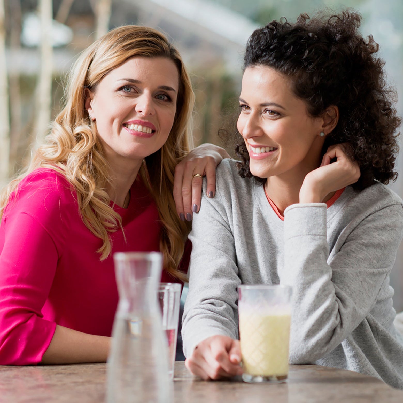Zwei Frauen sitzen zusammen, lächeln und plaudern über Abnehmtipps. Die eine genießt den Shake der Precon Probierwoche EU (7 Tage), während sie sich bei Getränken entspannen. Ein Glaskrug und Gläser auf dem Tisch unterstreichen den freundschaftlichen Moment.