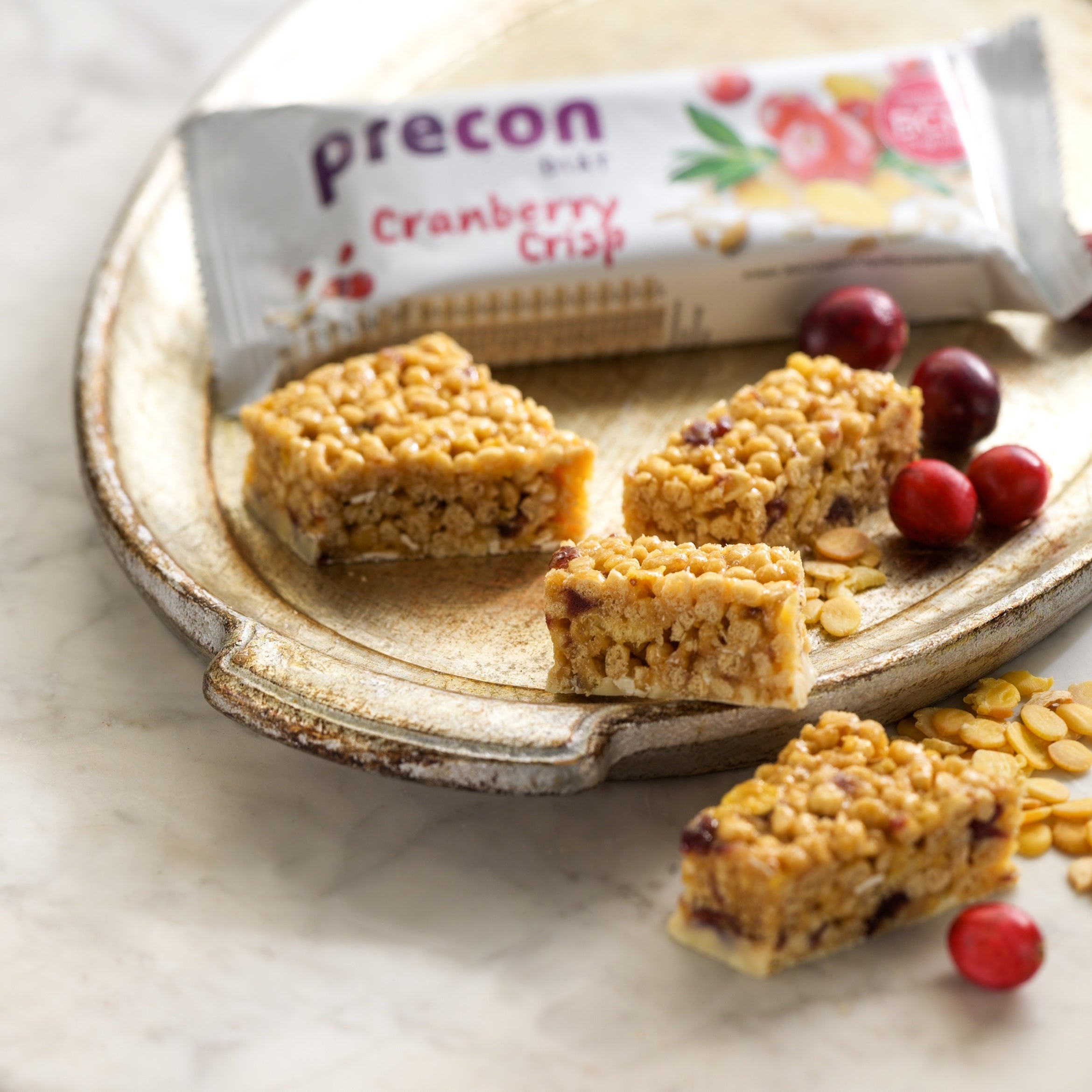 Auf einem Keramikteller liegen vier Stück Cranberry-Crisp-Müsliriegel, Cranberries und Kürbiskerne. Im Hintergrund ist ein verpackter Precon 3-Monats-Mega-Vorrats-Paket CH-Riegel auf einer hellen Marmorfläche teilweise sichtbar.