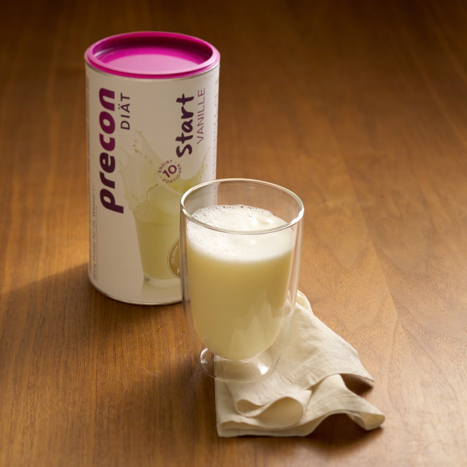 Ein Glas Diätdrink mit Vanillegeschmack steht auf einer beigen Serviette neben einem Behälter mit der Aufschrift Precon Start Shake Vanille - ideal für deinen Diätstart oder als leckerer Start Shake auf dem Weg zum Abnehmen.