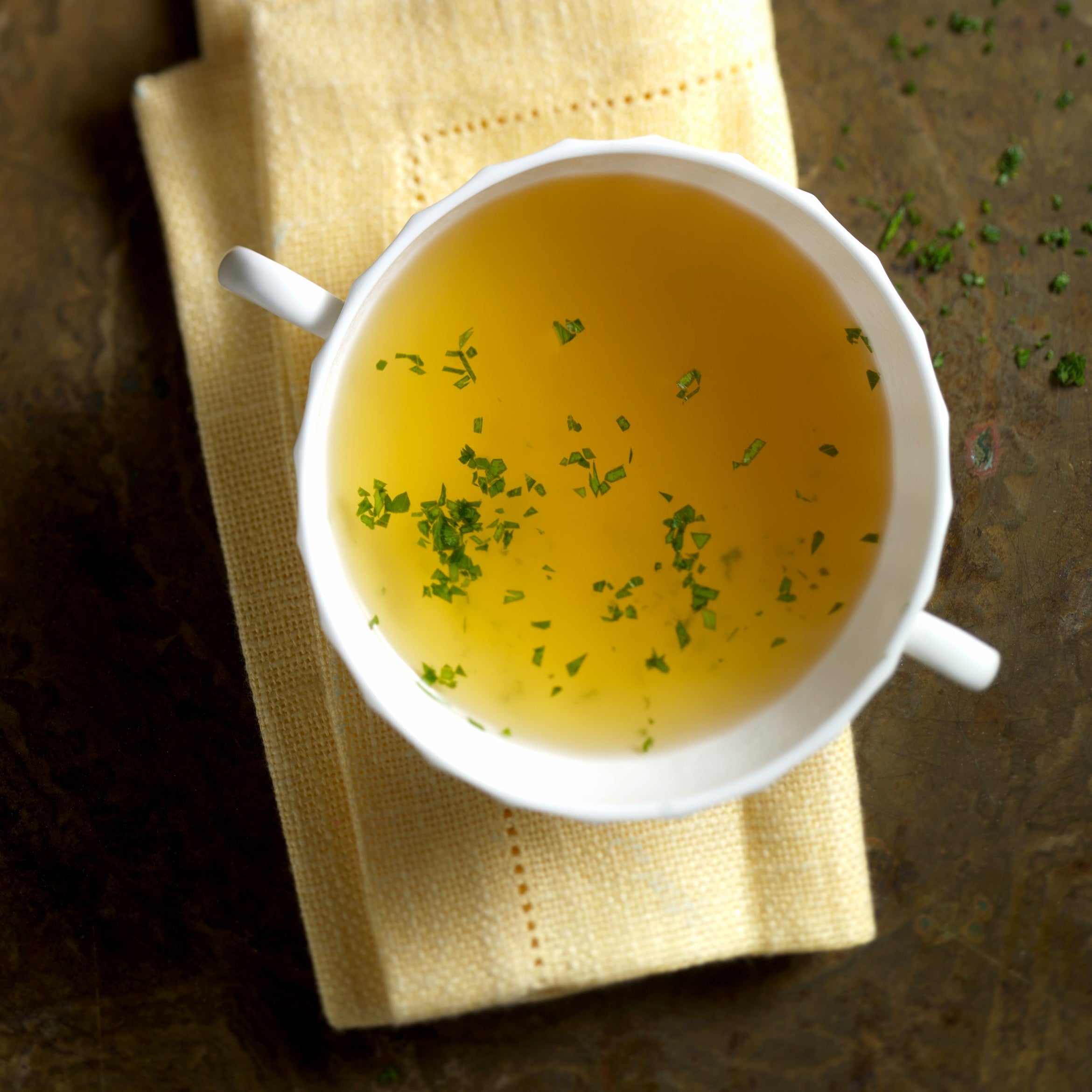 Eine weiße Schale mit Precon Special Kräuter, einer klaren Brühe, die ideal für Diät und Fettverbrennung ist, wird mit frischen Kräutern bestückt und auf einer gefalteten beigen Serviette auf einer rustikalen braunen Fläche angerichtet.