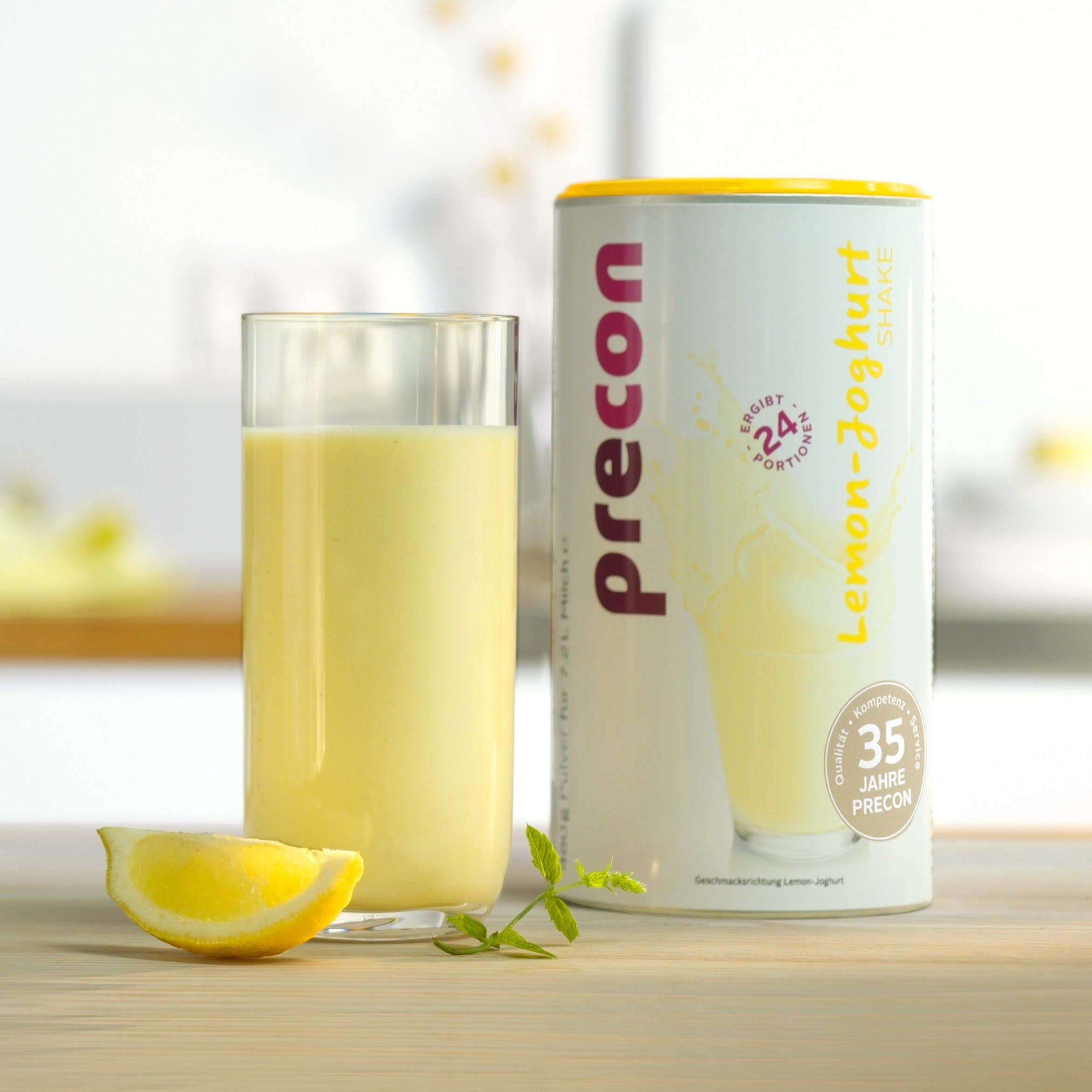 Ein Glas cremig-gelber Zitronen-Joghurt-Shake steht neben einer Zitronenspalte, frischer Minze und einer Probierwoche EU (7 Tage)-Box - und zeigt die leckeren Diät-Produkte von Precon für erfolgreiches Abnehmen.