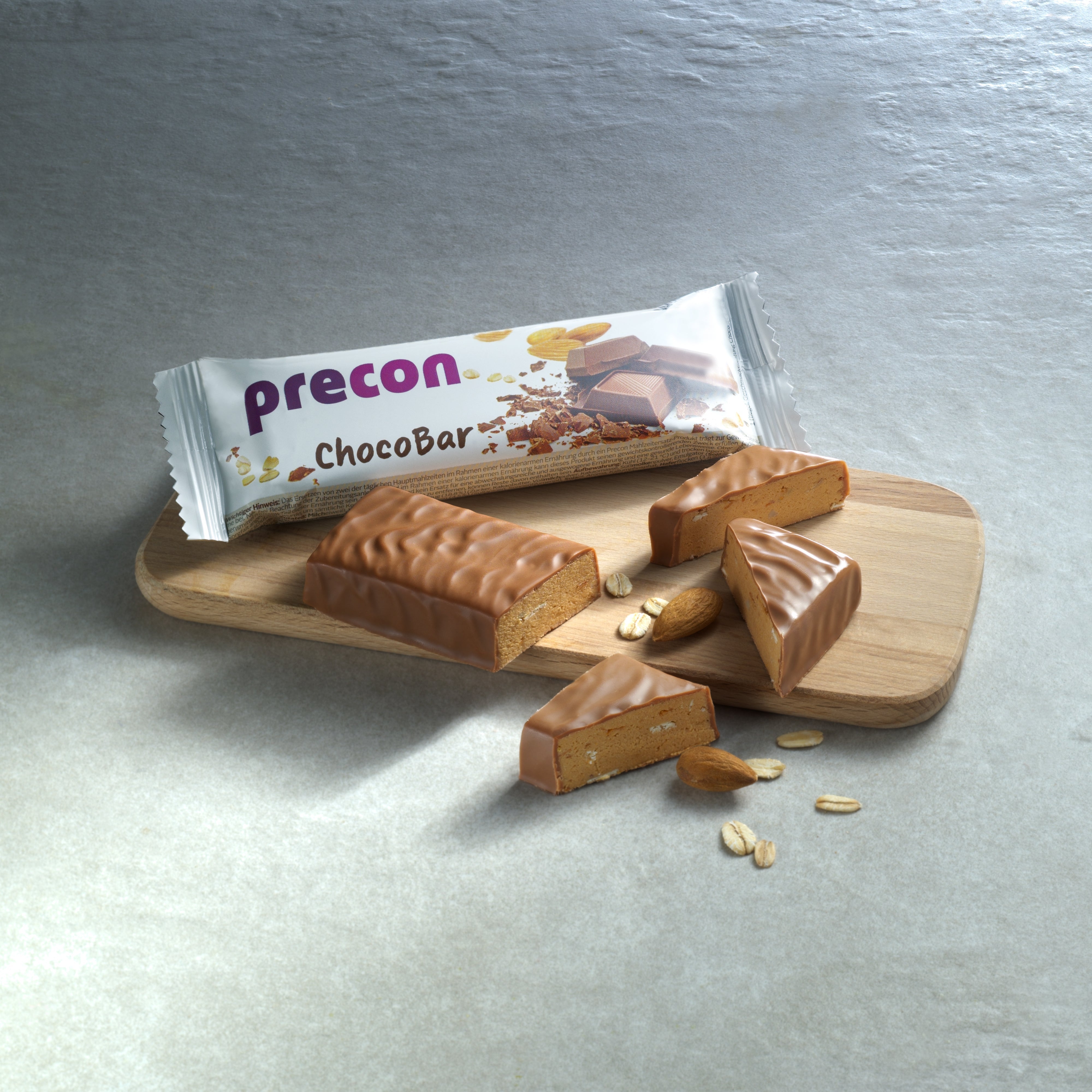 Ein teilweise ausgepackter Precon ChocoBar Riegel liegt auf einem Holzbrett mit Riegelstücken, Haferflocken und Nüssen - perfekt als leckerer Precon Diät Mahlzeitenersatz auf einer hellgrauen Fläche.